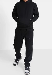 Sudadera negra con bolsillo frontal y puños de canalé, combinada con pantalones cargo negros que tienen múltiples bolsillos y cremalleras. Zapatillas negras.