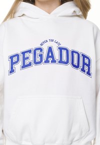 Sudadera blanca con un bolsillo frontal, con texto azul que dice "PEGADOR" y "NEVER TOO LATE" en un diseño de arco. Textura de tela suave.