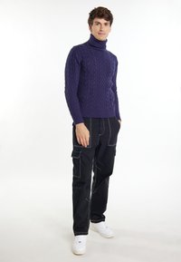 Maglione a collo alto a trama treccia viola abbinato a pantaloni cargo neri con cuciture bianche e tasche laterali, completato da sneakers bianche.