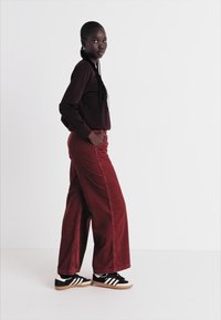 Pantaloni din catifea cord cu nuanță de vișiniu, cu buzunare laterale, asortați cu un top cu mânecă lungă de aceeași culoare și adidași negri cu dungi albe, pe un fundal alb.