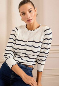 Maglione bianco con righe orizzontali blu navy e bordi ondulati, caratterizzato da un motivo a maglia testurizzato e dettagli dorati sulle spalle. Indossato con jeans blu.