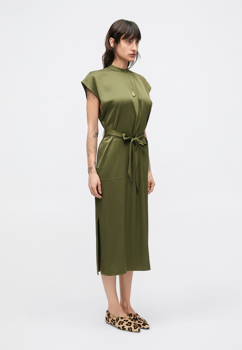 Olive green knee-length dress with cap sleeves, keyhole detail, tie waist, side slits, and two front pockets. Worn with leopard print shoes.