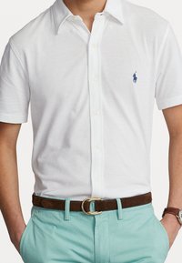 Polo shirt bianco a maniche corte con tessuto testurizzato, caratterizzato da un logo ricamato blu sul petto; abbinato a chinos azzurri chiari e una cintura marrone.