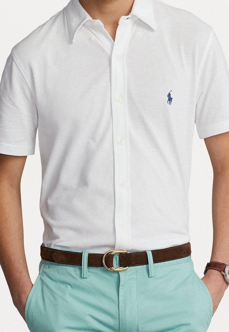 Polo shirt bianco a maniche corte con tessuto testurizzato, caratterizzato da un logo ricamato blu sul petto; abbinato a chinos azzurri chiari e una cintura marrone.