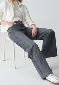 Persona sentada en una silla blanca, vistiendo una chaqueta blanca, una blusa clara, jeans anchos grises y zapatillas blancas con suelas de goma.