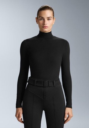 Crni dugi rukav s visokim ovratnikom, napravljen od glatkog materijala; uparen s hlačama visokog struka koje imaju strukturirani, teksturirani pojas.