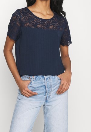 Vrouw draagt een navyblauw topje met korte mouwen en inzetstukken van kant, gecombineerd met lichtblauwe high-waisted jeans, handen in de voorzakken.