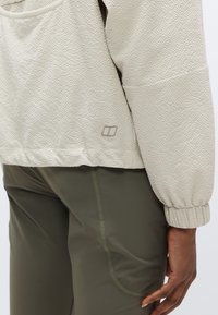 Veste beige clair texturée avec des poignets élastiques, présentant un petit logo sur le bas côté. Associée à un pantalon ajusté de couleur vert olive.