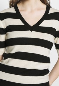 Žena oblečená v krátkém rukávu svetr s širokými vodorovnými pruhy v černé a béžové barvě s výstřihem do V.