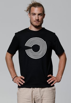 Zwarte t-shirt met een grafisch ontwerp van witte concentrische cirkels. Korte mouwen en een ronde hals, gemaakt van zachte stof. Model staat met handen op de heupen.