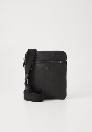 Sac bandoulière en cuir noir avec une poche zippée à l'avant et une sangle large en tissu ajustable sur un fond clair uni.