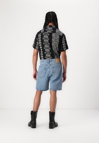 Cămașă neagră cu mâneci scurte, cu modele florale albe, asociată cu pantaloni scurți din denim albastru deschis și ghete negre până la gleznă, văzută din spate.