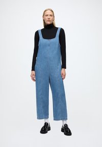 Lyseblå denimoverall med firkantet halsutringning, vide ben og sidelommer, kombinert med en svart høyhalset genser og svarte loafers.