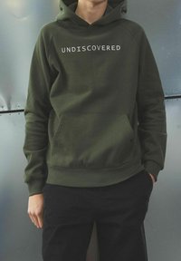 Oliwkowa bluza z kapturem z białym napisem "UNDISCOVERED", kangurzą kieszenią, prążkowanymi mankietami i dolnym ściągaczem, noszona z czarnymi spodniami na metalicznym tle.