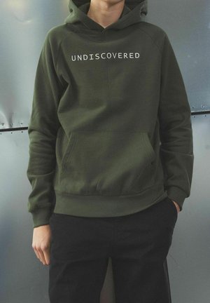 Hoodie vert olive avec le texte "UNDISCOVERED" en blanc, poche kangourou, poignets et ourlet côtelés, porté avec un pantalon noir sur un fond métallique.