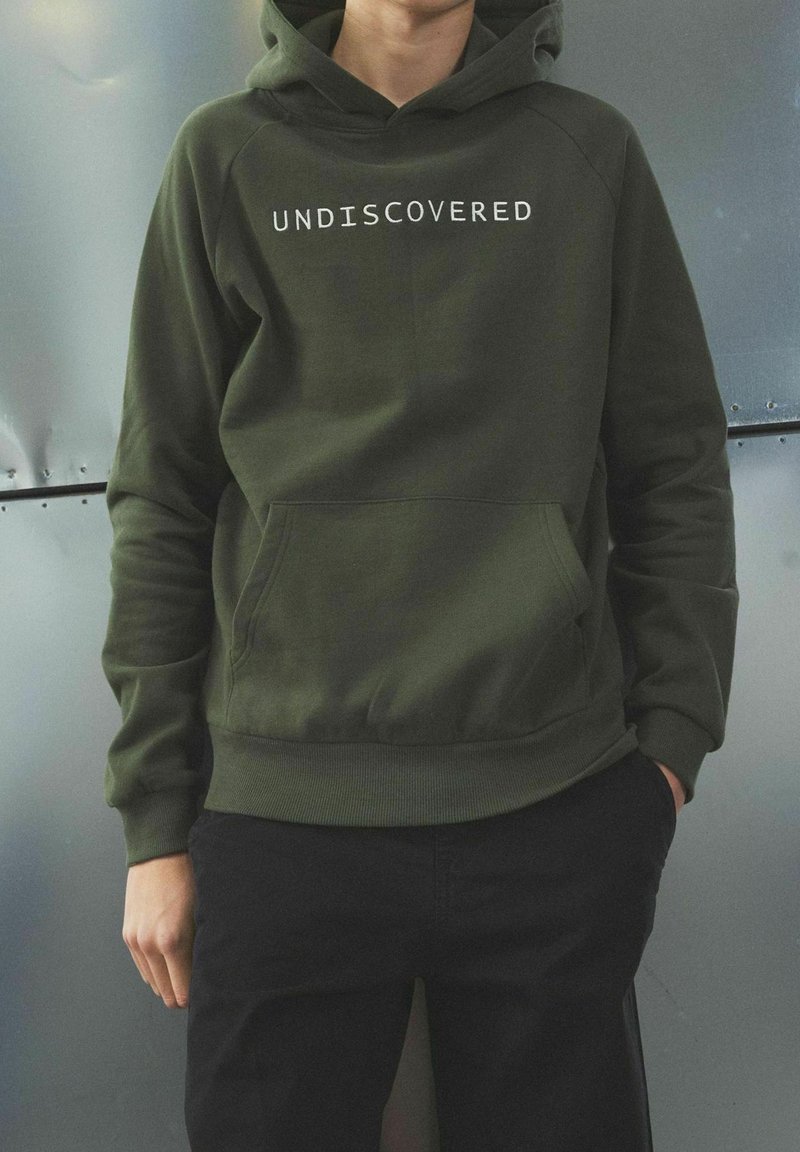 Oliwkowa bluza z kapturem z białym napisem "UNDISCOVERED", kangurzą kieszenią, prążkowanymi mankietami i dolnym ściągaczem, noszona z czarnymi spodniami na metalicznym tle.