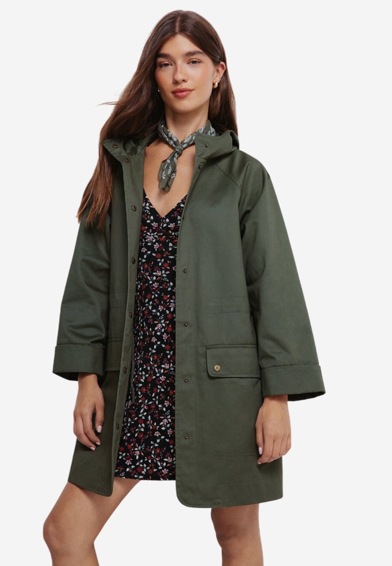 Olivengrøn, mellemlang regnjakke med hætte, trykknapper og to for lommer. Stylet over en sort blomsterkjole med korte ærmer.