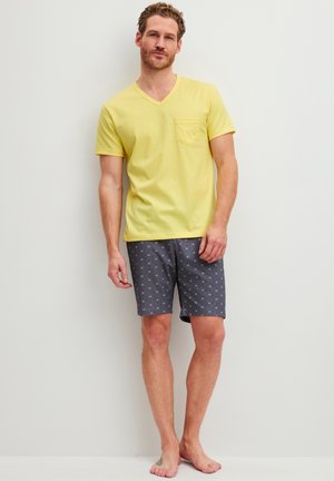 Man staat blootsvoets, draagt een geel T-shirt met korte mouwen en V-hals en donkere korte broek met patroon, tegen een effen lichte muur.