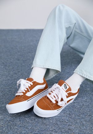 Person iført hvide sokker og lyseblå jeans med brune og hvide Vans sneakers, siddende på et blåt tæppe.