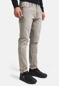 Jeans de mezclilla gris deslavados con detalles desgastados y múltiples roturas, combinados con zapatillas negras de diseño bajo y cordones planos.