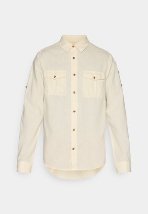 Chemise longue à manches beiges claires en tissu au toucher doux, dotée de deux poches avant et de boutons dorés. Conception à ourlet arrondi.
