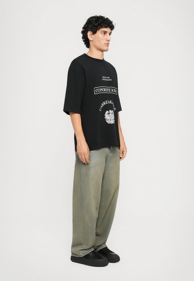 Ung mann står på siden iført en svart grafisk T-skjorte, løse falmede grønne jeans og svarte sneakers på en enkel hvit bakgrunn.