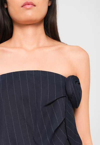 Vrouw in een strapless marineblauwe pinstripe top met een opgevouwen stofdetail aan één kant, waarbij de blote schouders en hals zichtbaar zijn.