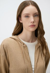 Beige zip-up hoodie met een capuchon, ribbeltextuur en zichtbare stiksels. Gedragen over een wit shirt, waardoor het zachte materiaal wordt benadrukt.