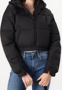 Czarna pikowana kurtka z kapturem, z zamek błyskawicznym z przodu, elastycznymi mankietami i skróconym krojem, zestawiona z niebieskimi dżinsami.