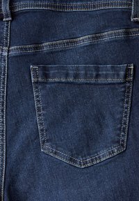 Tummansiniset farkut, joissa on suorakaiteen muotoinen takatasku hienovaraisella tekstuurilla ja kontrastivärisellä keltaisella ompeleella saumoissa.