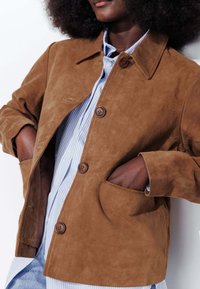 Veste en daim marron avec un col, deux poches avant et trois boutons, portée sur une chemise rayée bleu clair et blanche.