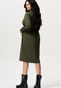 Olivgrünes Hemdkleid mit langen Ärmeln, tailliertem Bund und knielanger Länge. Kombiniert mit schwarzen Knöchelschuhen mit Schnallen.