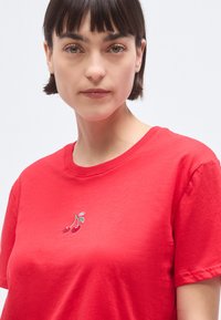 T-shirt en coton rouge à manches courtes et col rond, avec un petit motif de cerise brodé en vert et rouge sur la poitrine.