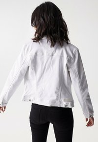 Chaqueta de mezclilla blanca con mangas largas y un diseño ligeramente ajustado, que presenta una etiqueta en la parte baja de la espalda y detalles de metal.