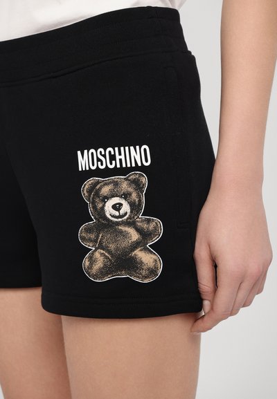 Juodi medvilniniai šortai su rudu lokio grafiku su šešėliu ir žodžiu "MOSCHINO", atspausdintu ryškiu baltu šriftu viršuje.