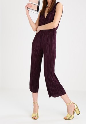 Kvinde iført en ærmeløs, dyb blommefarvet plisseret jumpsuit, der holder en lille blush- og sort clutch samt guldåbne tåblokhælsandaler.
