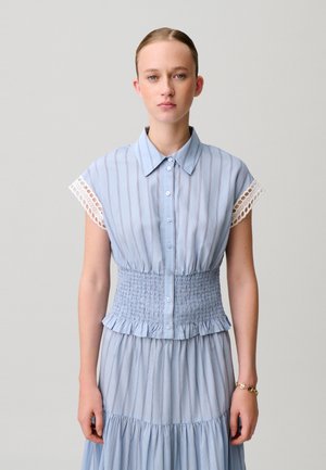 Jeune femme portant une robe chemise rayée bleu clair à taille smockée et manches courtes bordées de dentelle, debout devant un fond uni.