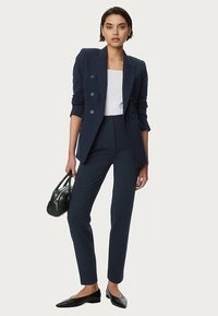 Marineblauwe getailleerde blazer met dubbele rij knopen, wit topje, slim-fit broek, zwarte handtas en zwarte puntige platte schoenen.