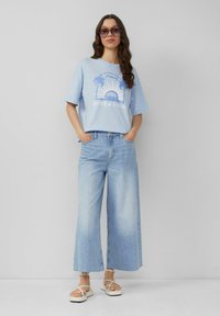 Lichtblauwe t-shirt met korte mouwen en grafisch ontwerp, gecombineerd met wijde lichtblauwe jeans en bandjesandalen, tentoongesteld tegen een neutrale achtergrond.