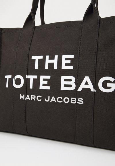 Crna platnena torba s debelim bijelim natpisima koji glase "THE TOTE BAG" i "MARC JACOBS." Ima čvrste ručke i strukturalni oblik.