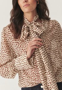 Femme portant un blouse beige avec des pois marron, des manches longues bouffantes et un grand noeud noué au cou, ajustant le noeud avec sa main.
