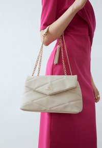 Beige prošívaná kabelka se zlatým logem, s klopovým uzávěrem a odnímatelným řetízkem, který má detail s třásněmi.