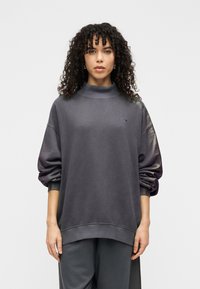 Γκρι υπερμεγέθη φούτερ με ψηλό γιακά, μανσέτες με ριμπ ύφανση και γυαλιστερές λεπτομέρειες στα μανίκια. Διαθέτει μικρό κεντητό λογότυπο στο στήθος.