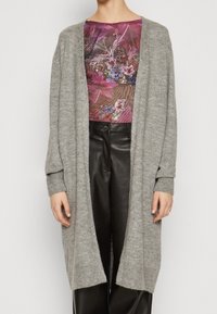 Šedý pletený cardigan s volným střihem a dlouhými rukávy, s žebrovanými manžetami a otevřeným předním designem, nošený přes vzorované tričko.