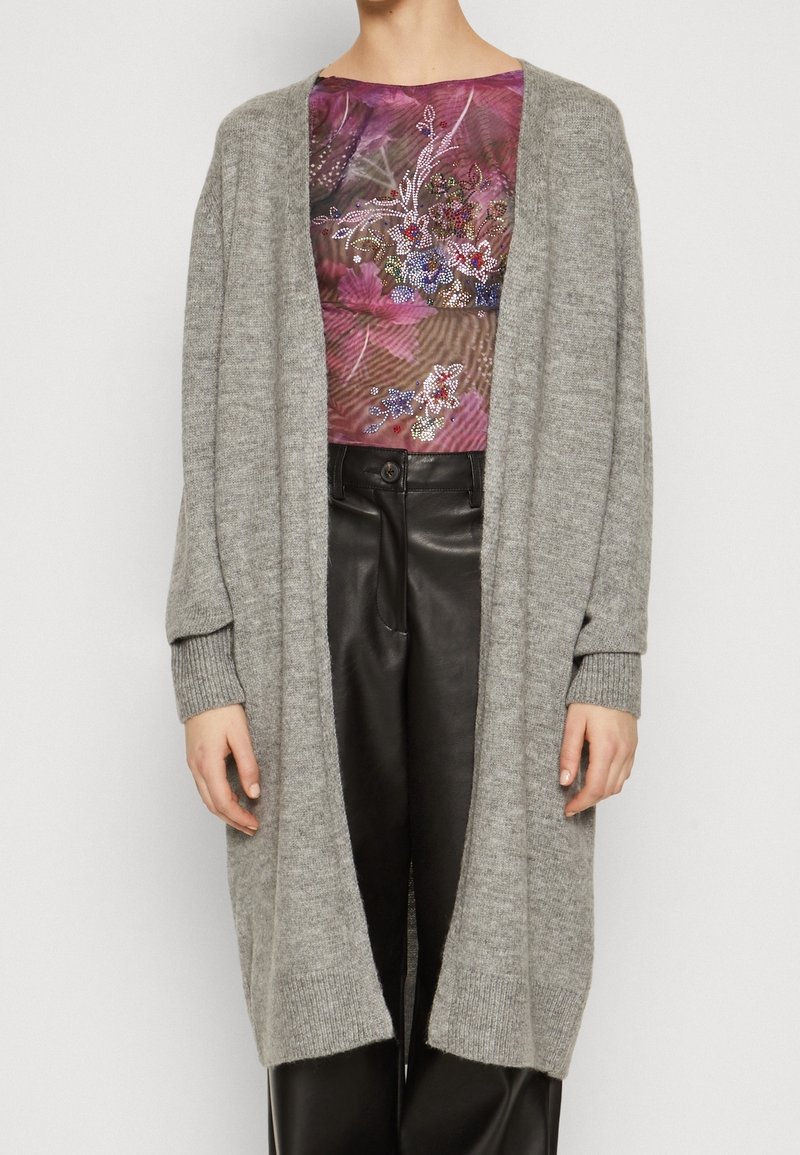 Šedý pletený cardigan s volným střihem a dlouhými rukávy, s žebrovanými manžetami a otevřeným předním designem, nošený přes vzorované tričko.