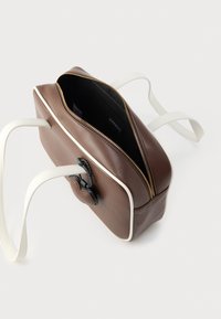 Καφέ δερμάτινη τσάντα με λευκή επένδυση και λουριά, κλείσιμο με φερμουάρ και υφή μαύρου διακοσμητικού στη μία πλευρά. Το εσωτερικό είναι μαύρο.