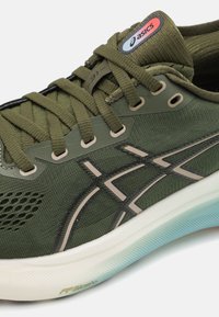 Olivenfargede treningssko med mesh-overdel, polstret krage og svarte detaljer. Har Asics-logo og teksturert såle med lyseblå gradient.