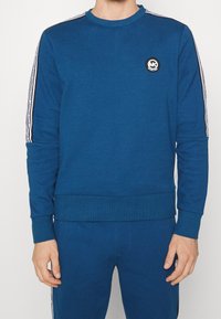 Felpa blu in morbido tessuto con colletto rotondo, polsini e orlo a coste. Presenta strisce laterali bianche e un logo circolare sul petto.