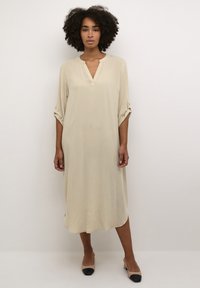 Beige midi šaty s výstřihem do V, srolovanými rukávy zabezpečenými knoflíky a hladkou texturou. Spárované s béžovými a černými dvoutónovými botami.
