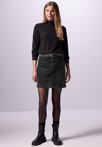 Pull en col roulé noir, mini-jupe en velours côtelé vert avec passants de ceinture, poches avant, et bottines noires, accompagnées de collants noirs.